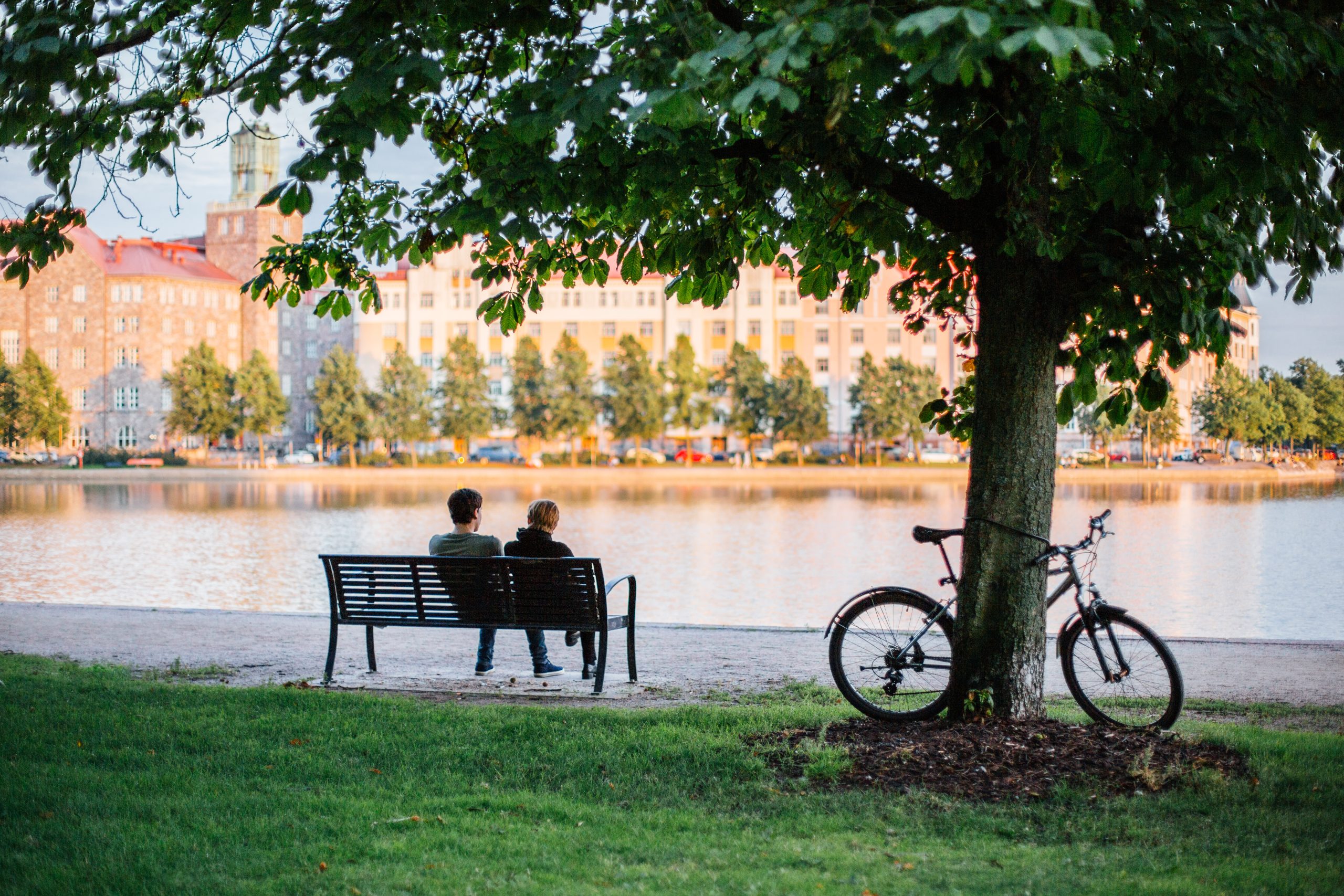 Two people sitting on a bench looking at views over Eläintarhanlahti, with a bike resting against a tree